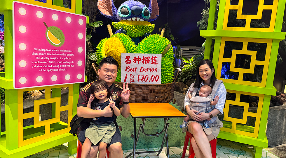 Family posing beside a durian-themed display and floral Stitch sculpture at an indoor exhibit